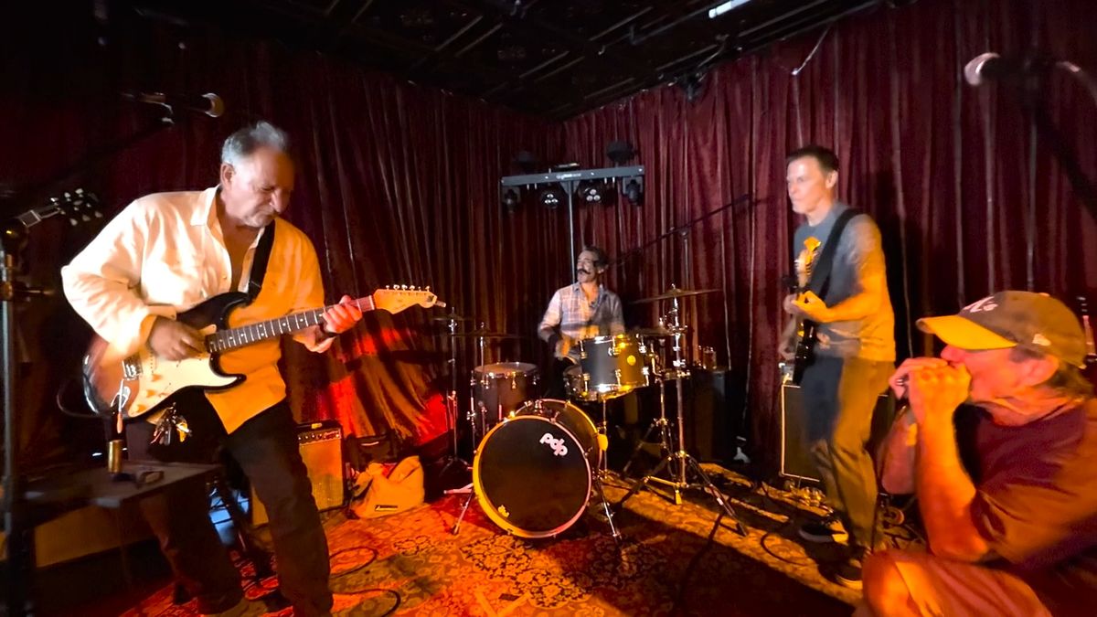 The Boogie Chillun Band on stage at Mac's — guitarist, drummer and bassist under red stage curtains, with Catfish Jack playing harmonica at the right of frame