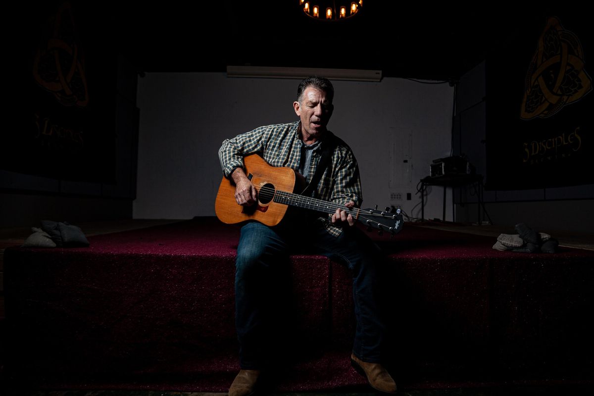 Catfish Jack sitting on the edge of a stage, singing and playing acoustic guitar under a single spotlight