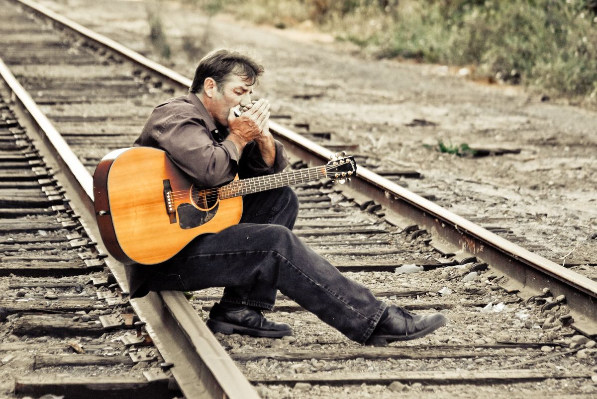 Catfish Jack sitting on railroad tracks with an acoustic guitar across his lap, playing harmonica