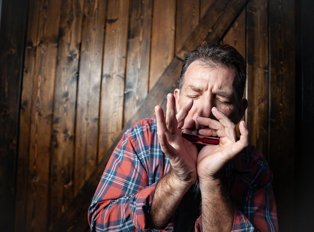 Catfish Jack playing blues harmonica, both hands cupped around the harp and his eyes closed, against a dark barn-wood wall