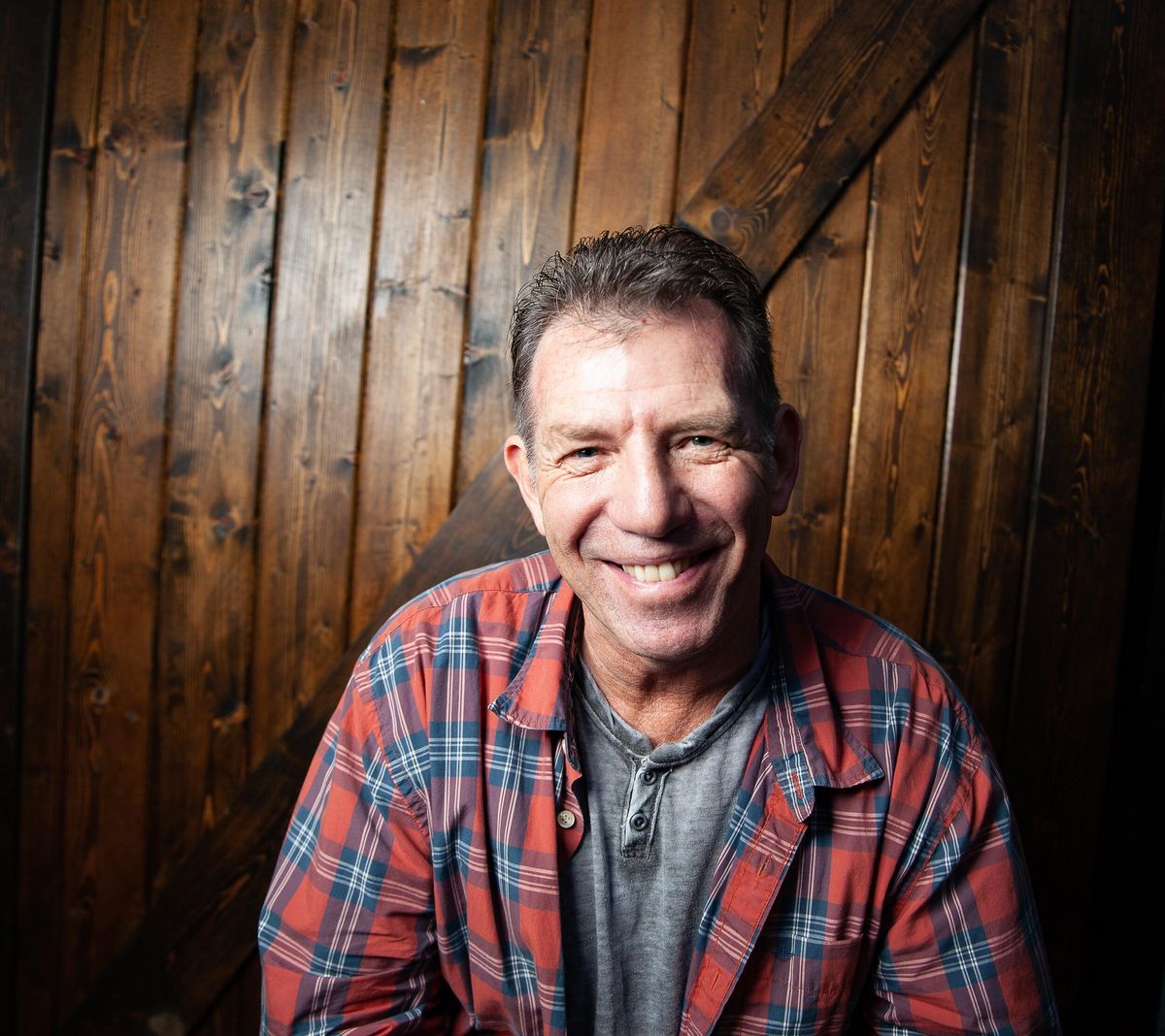 Catfish Jack smiling in a red plaid shirt against a dark barn-wood wall