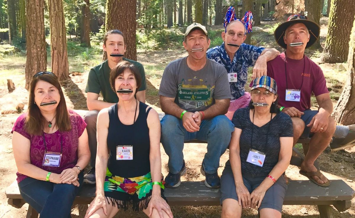 Seven adult harmonica students posing in a redwood grove with harmonicas held in their mouths, name badges on
