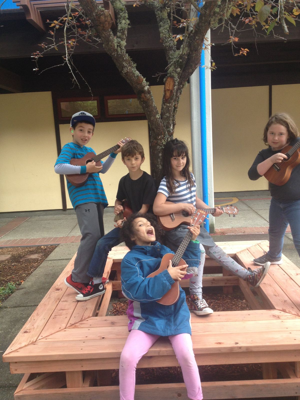Five kids playing ukuleles around a picnic table outside a school, one belting out the song